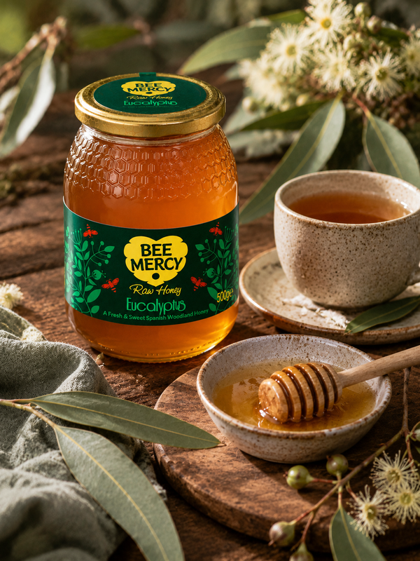 A jar of Bee Mercy Raw Honey Eucalyptus on a rustic wooden table, styled with a ceramic cup of tea, a honey dipper in a small bowl, and fresh eucalyptus leaves and flowers.