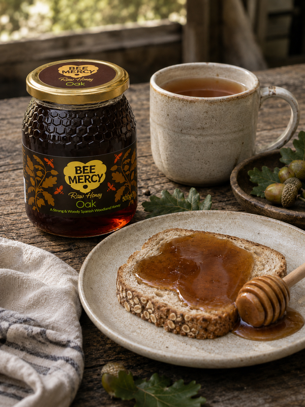 A jar of Bee Mercy Rose Honey Oak on a rustic wooden table next to a cup of tea, a slice of bread with honey on a plate, and an acorn.