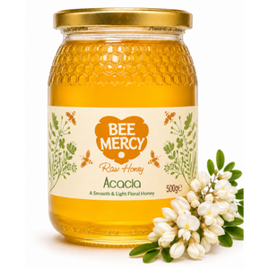 A 500g glass jar of Bee Mercy Raw Honey with Acacia flavor, featuring a gold lid and a cream-colored label with green botanical illustrations. A small cluster of white acacia flowers is placed next to the jar on a clean white background.
