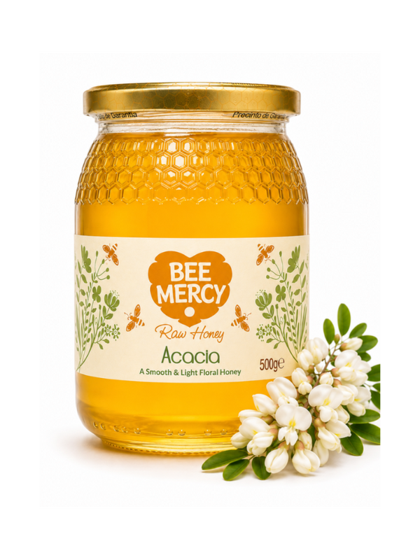 A 500g glass jar of Bee Mercy Raw Honey with Acacia flavor, featuring a gold lid and a cream-colored label with green botanical illustrations. A small cluster of white acacia flowers is placed next to the jar on a clean white background.
