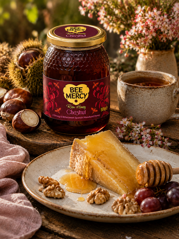 A jar of Bee Mercy Raw Honey Chestnut placed on a rustic wooden table beside a plate with honey, walnuts, grapes, and a honey dipper. A cup, flowers, and chestnuts complete the styled scene.