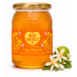 A 500g glass jar of Bee Mercy Raw Honey Citrus Blossom on a white background, styled with a green lime and white citrus blossoms.