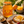 A jar of Bee Mercy Citrus Blossom honey sits on a rustic wooden table surrounded by food props, including a plate with toast topped with honey and orange slices, fresh berries, a honey dipper, oranges, and small bowls of flowers and blossoms.