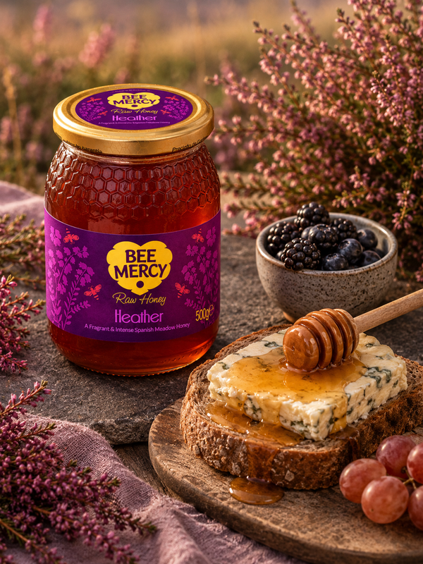 A jar of Bee Mercy Heather raw honey displayed on a rustic stone surface with a slice of blue cheese, honey dipper, grapes, and a small bowl of blackberries, surrounded by heather flowers in an outdoor field.