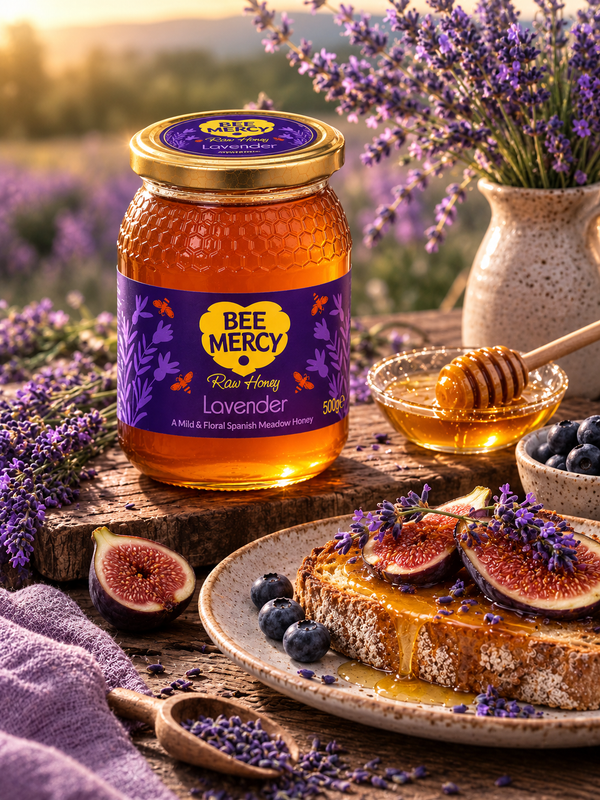 A jar of Bee Mercy Lavender Raw Honey displayed on a rustic wooden table with a plate of honey toast topped with figs and lavender, alongside a honey dipper, blueberries, and a vase of lavender in a warm, sunlit outdoor setting.