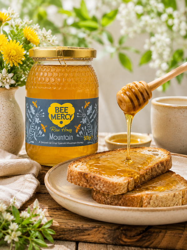 A jar of BEE MERCY Raw Honey Mountain honey sits on a rustic wooden table next to a plate of toast with honey dripping from a wooden dipper, with flowers and a mug in the background.
