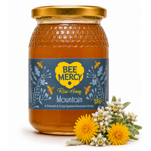A 500g glass jar of Bee Mercy Raw Honey Mountain honey, featuring a blue and yellow label, a gold lid, and fresh yellow flowers and white wildflowers placed in front on a white background.