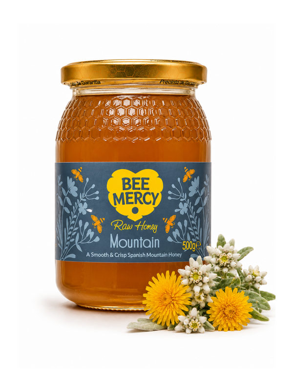 A 500g glass jar of Bee Mercy Raw Honey Mountain honey, featuring a blue and yellow label, a gold lid, and fresh yellow flowers and white wildflowers placed in front on a white background.