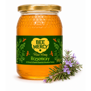 A 500g glass jar of Bee Mercy Raw Honey Rosemary, featuring a green and yellow label, a gold lid, and a sprig of fresh rosemary on a white background.