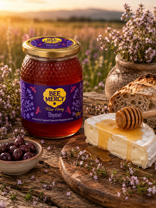 A jar of Bee Mercy Thyme Honey is displayed on a rustic wooden table alongside a slice of Brie cheese with honey drizzle, a honey dipper, olives, and lavender flowers, set against a blurred field at sunset.
