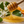 A wooden honey dipper filled with thick golden honey, with honey dripping into a pool below. The scene includes green leaves, white blossoms, and orange slices in a softly blurred natural setting.
