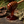 A wooden honey dipper filled with glossy amber honey, with honey dripping down, set on a dark stone surface with pine needles, bark, and leaves around it.