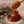 Glossy amber honey pooling on a stone surface, with a wooden honey dipper lifting some of the honey above it. Pink heather flowers and green stems are placed along the left side of the scene.