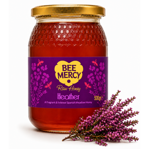 A 500g glass jar of Bee Mercy Raw Honey with a purple and yellow label, decorated with a sprig of purple heather on a white background.
