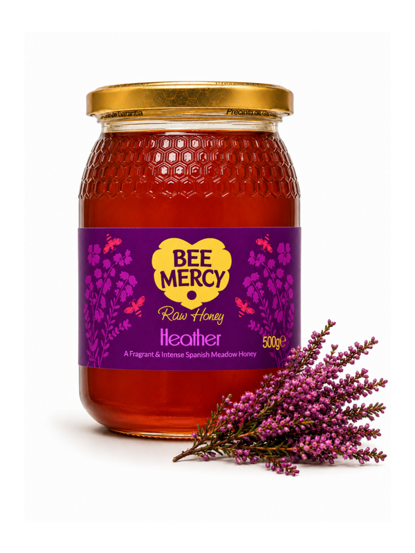 A 500g glass jar of Bee Mercy Raw Honey with a purple and yellow label, decorated with a sprig of purple heather on a white background.