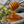 A wooden honey dipper filled with thick golden honey, with honey dripping onto a stone surface and surrounded by small wildflowers.